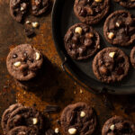 A plate holds a bunch of double chocolate chip macadamia nut cookies on a brown table.