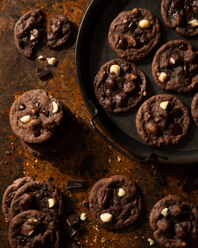 A plate holds a bunch of double chocolate chip macadamia nut cookies on a brown table.