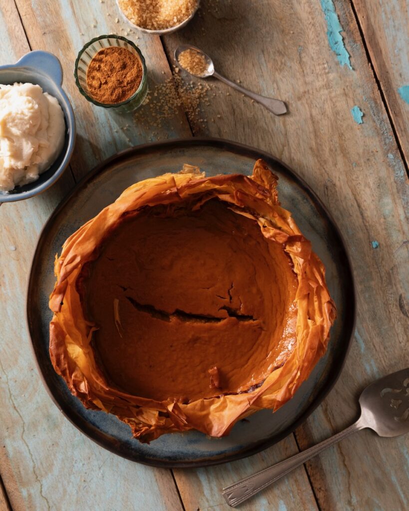 A pumpkin pie with a phyllo crust pits in a pan on a picnic table with small bowls of spices and spoons sitting around it.