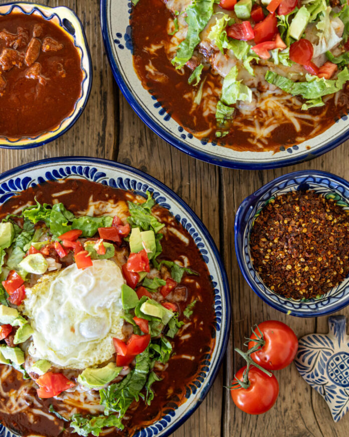 Freshly made Red Chile and Blue Corn Enchiladas by Carmella Padilla sit on a table with a side of beans, salsa, and tomatoes nearby.