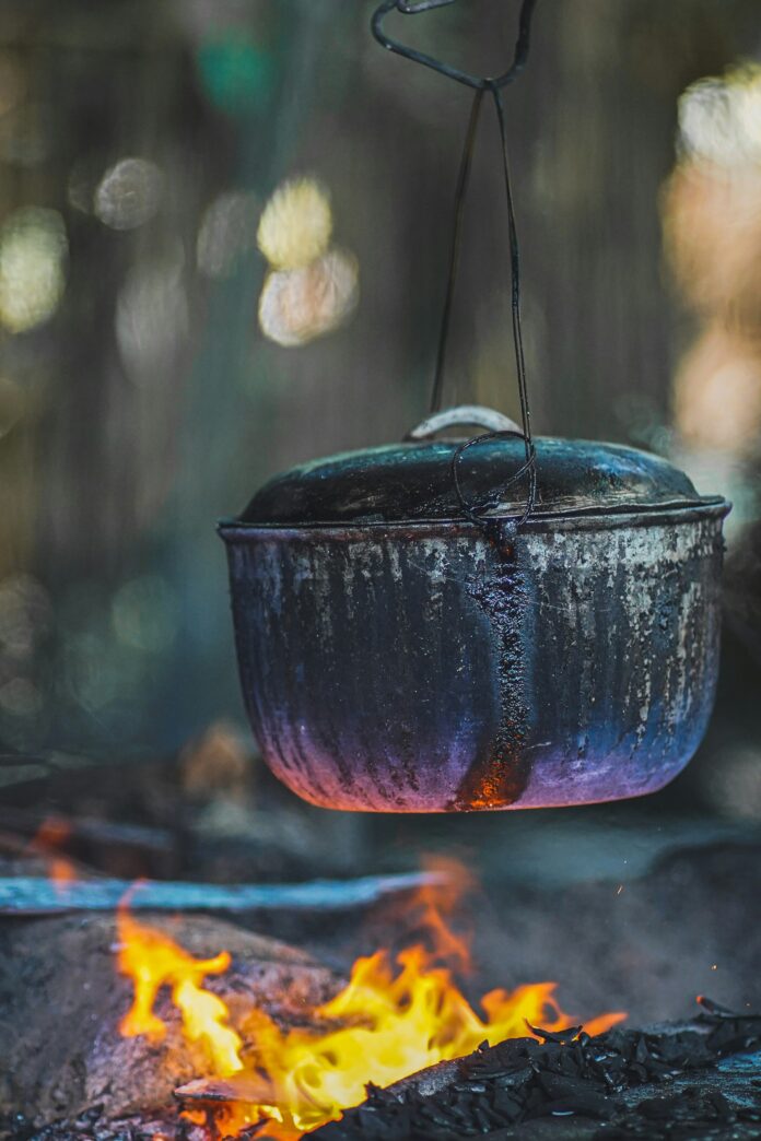 A dutch oven is held over an open campfire flame by a chain.