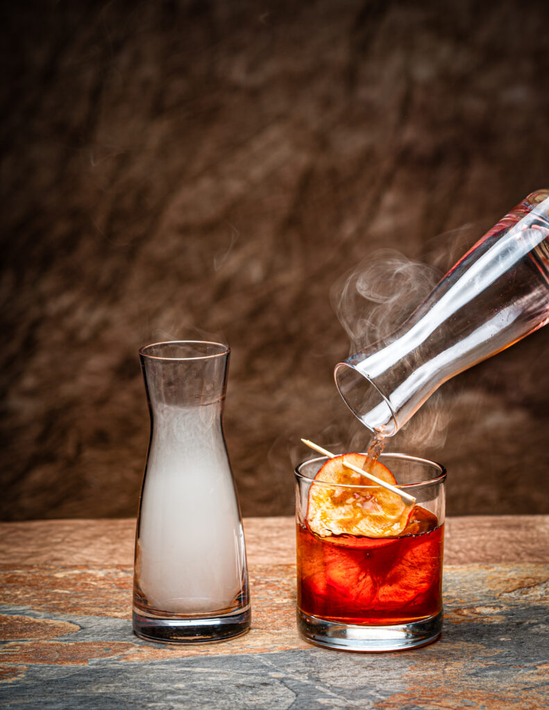 A short cocktail glass holds a red cocktail with an orange garnish next to a milk glass of smoke.