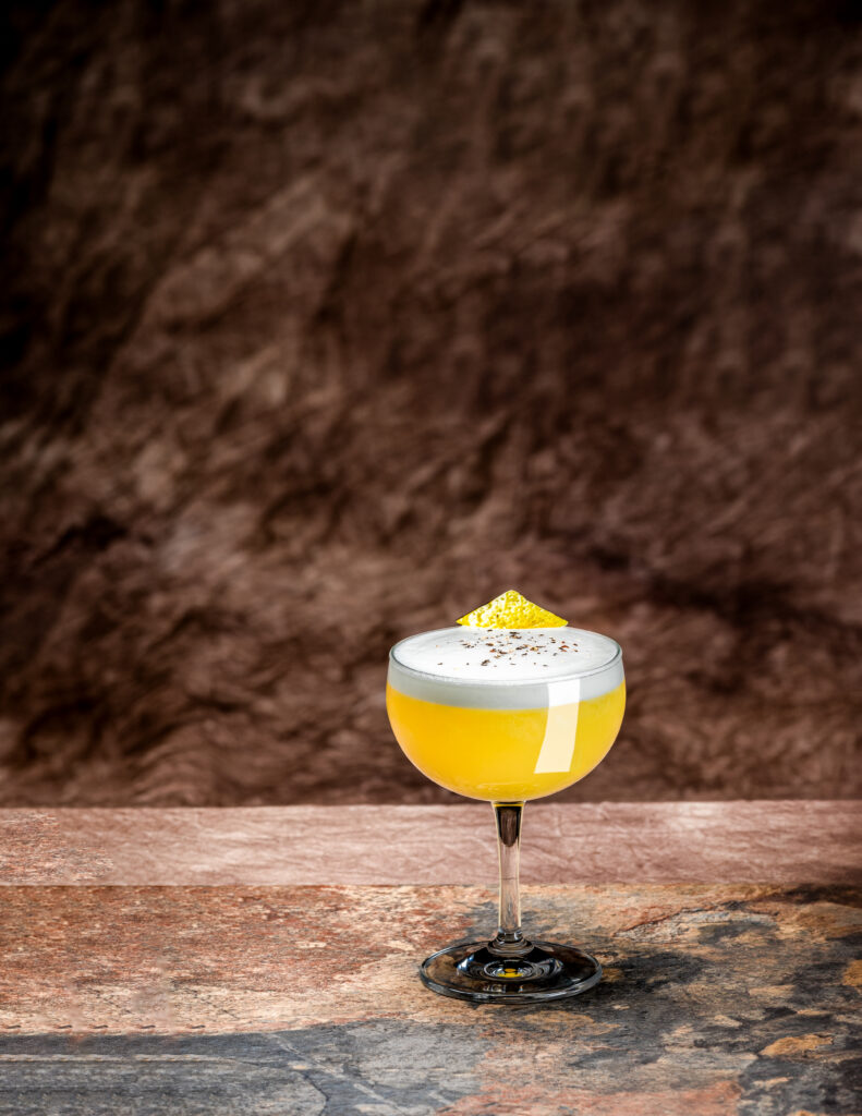 A rounded cocktail glass is filled with a yellow liquid and topped with a white egg foam and lemon peel. It sits on a brown table and background.