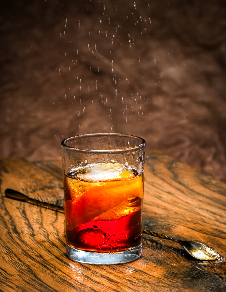 A shot glass holds a negroni cocktail with a stir stick sitting behind it on a wooden table.
