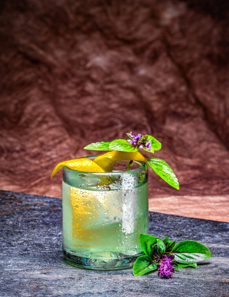 A short green cocktail glass holds a clear liquid garnished with lemon peels and flowers.