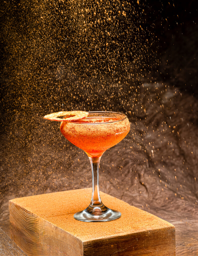 A tall cocktail glass holds an orange drink with a dried ring garnish and dusty brown sand falling overtop of it.