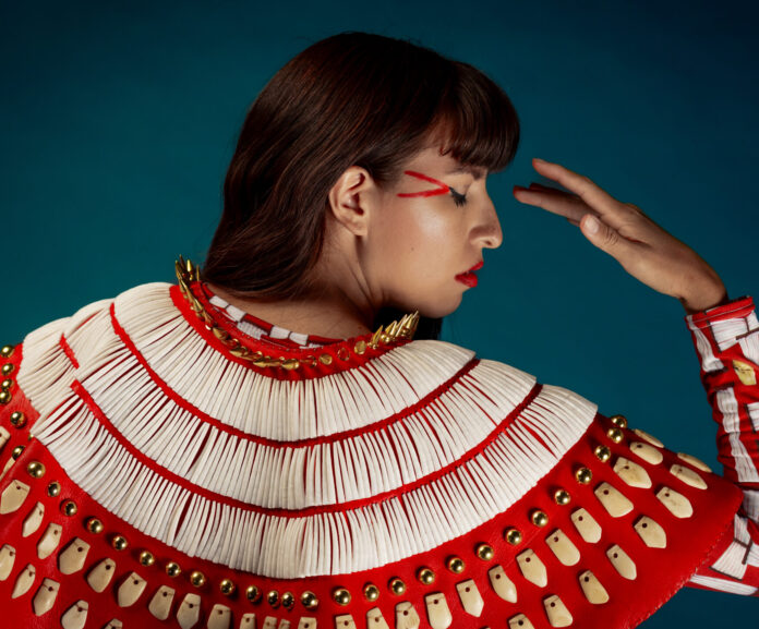 A woman in traditional red native american dress and makeup faces away from the camera, her head looking off to the side and her eyes closed.