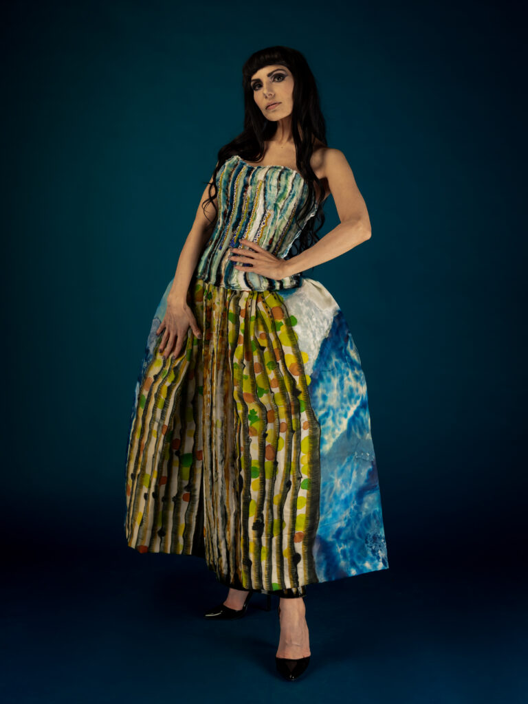 A woman poses in a multi colored and patterned dress against a blue background for SWAIA Native Fashion Week in Santa Fe