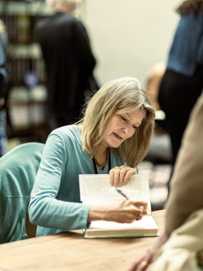 A woman in a blue long sleeve shirt signs the inside of a book on a wooden table.
