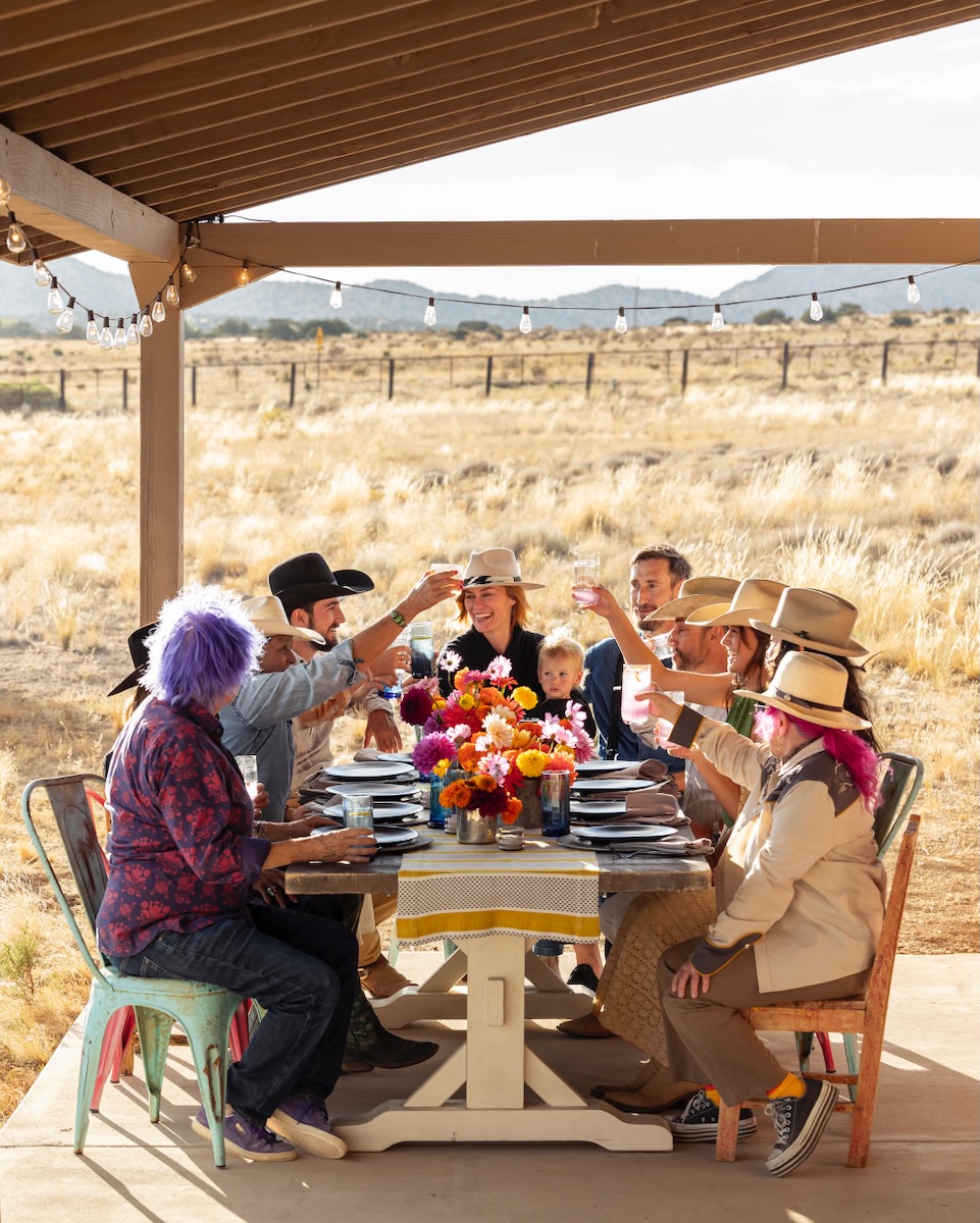 A family gathers at a wood table outside Double DD Ranch, each holding up their arm and drink to cheers as a ranch setting sits in the background.