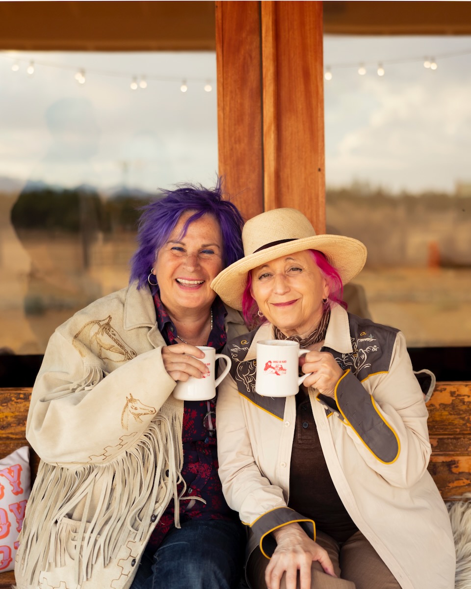 Two women who own Double DD Ranch sit on a bench and hold coffee mugs with long jackets on, one with purple hair and the other with pink hair and a hat.