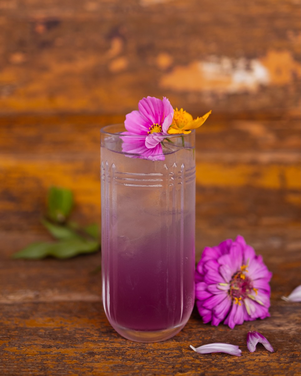 A cocktail with a purple gradient sits on a wooden table with a purple flower on top and one beside the glass as well.