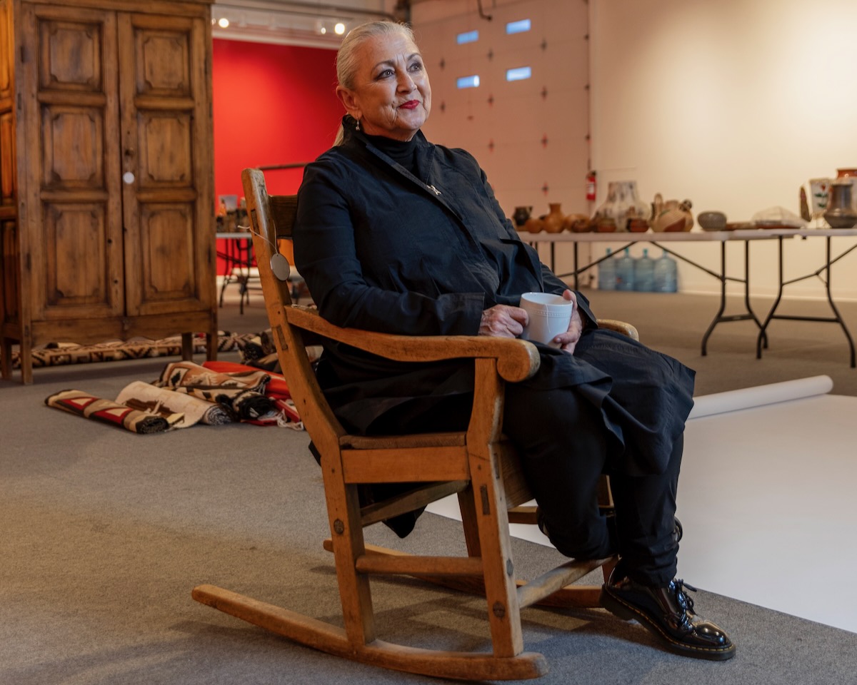 Gillian Blitch sits in the Santa Fe Art Auction house in a wooden rocking chair with a red wall behind her.