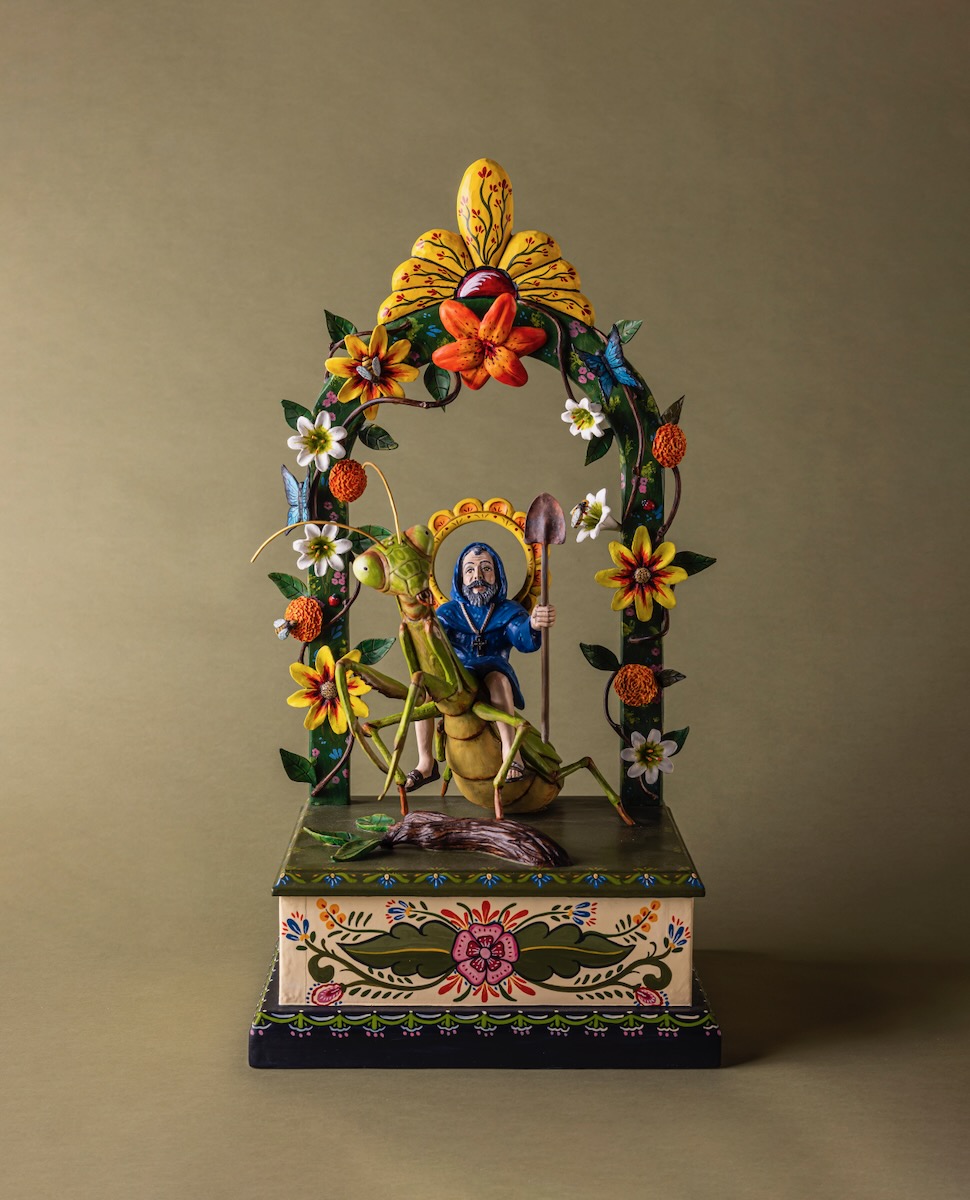 A wooden sculpture of San Fiacre under an arch of flowers from the Spanish Colonial Society sits against a brown background.