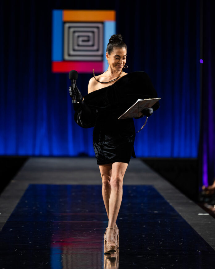 Amber Dawn Bear Robe stands on the SWAIA Native Fashion Week runway, clipboard and microphone in hand with the SWAIA logo behind her.