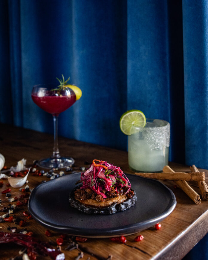 A blue plate holds a House-Chorizo Memela while a tall red cocktail sits to the back left and a short pale cocktail with a lime wheel sits to the back right.