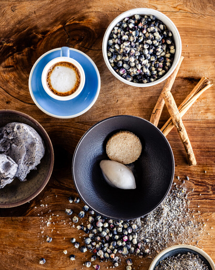 A small blue bowl holds two scoops of Blue Corn Gelato on top of cookies with another bowl of it to the left. A latte and blue flowers sit above the bowls.