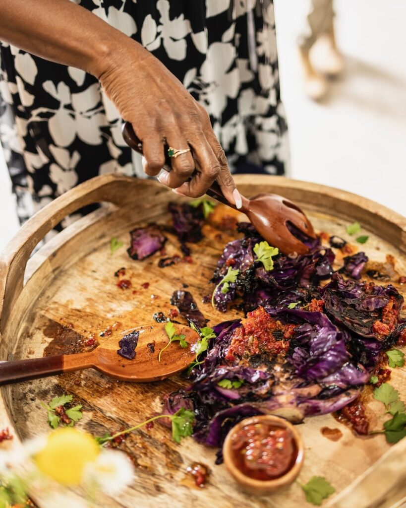 A basket platter holds a variety of charred red cabbage as a woman uses utensils to serve the dish.