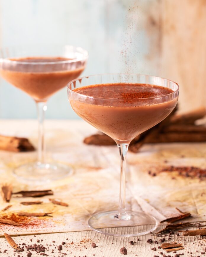 Two cocktail glasses hold a mole chocolate cocktail on a wood table with powdered chocolate surrounding them.