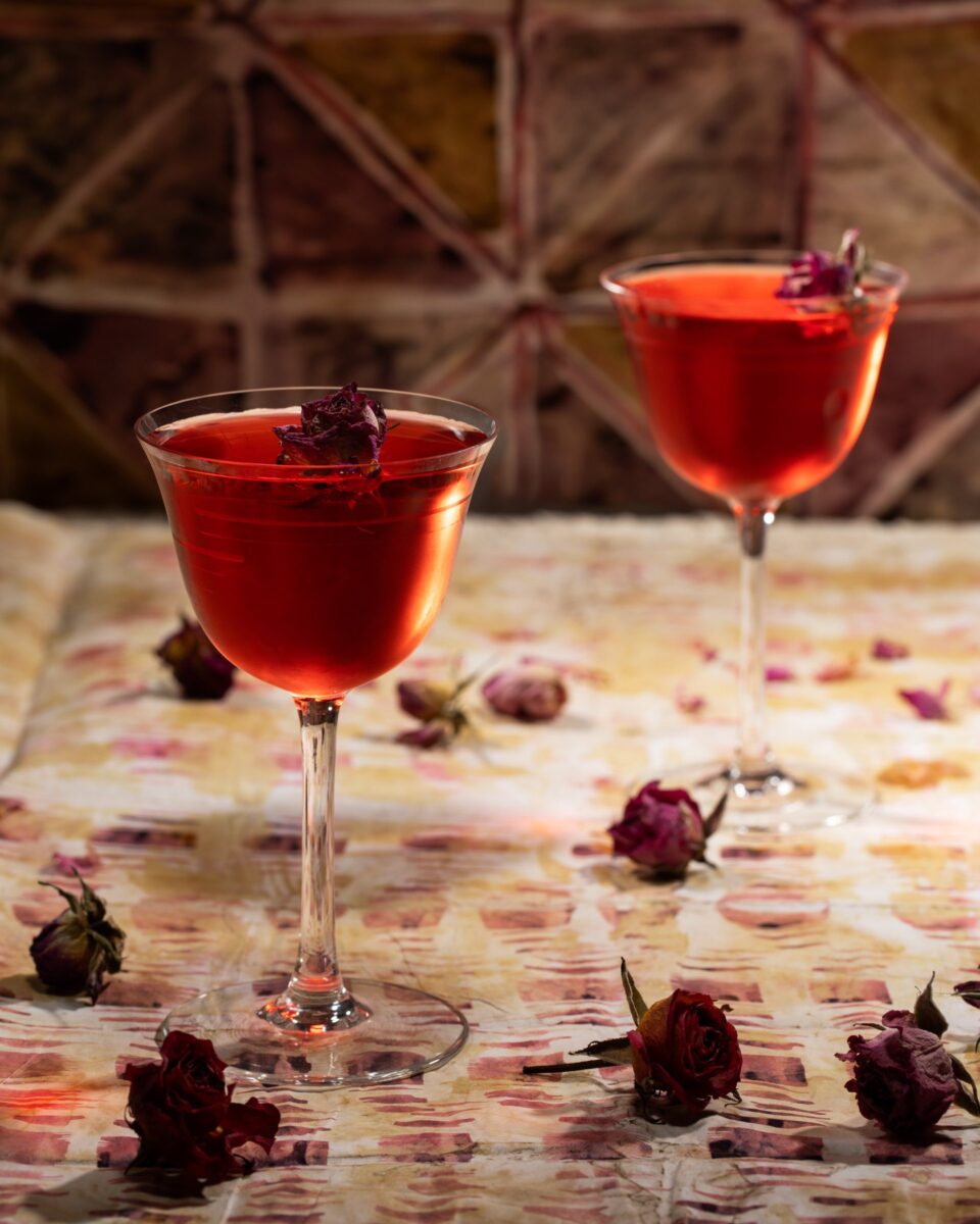 Two tall cocktail glasses with a red liquid inside are topped with dried rose buds as other dried rose buds sit on a tablecloth below the glasses.