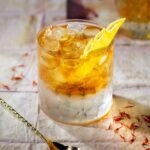 A cocktail glass holds a brown liquid for the I Feel a Sin Coming On Cocktail garnished with a lemon peel with a bar spoon sitting in front of it.