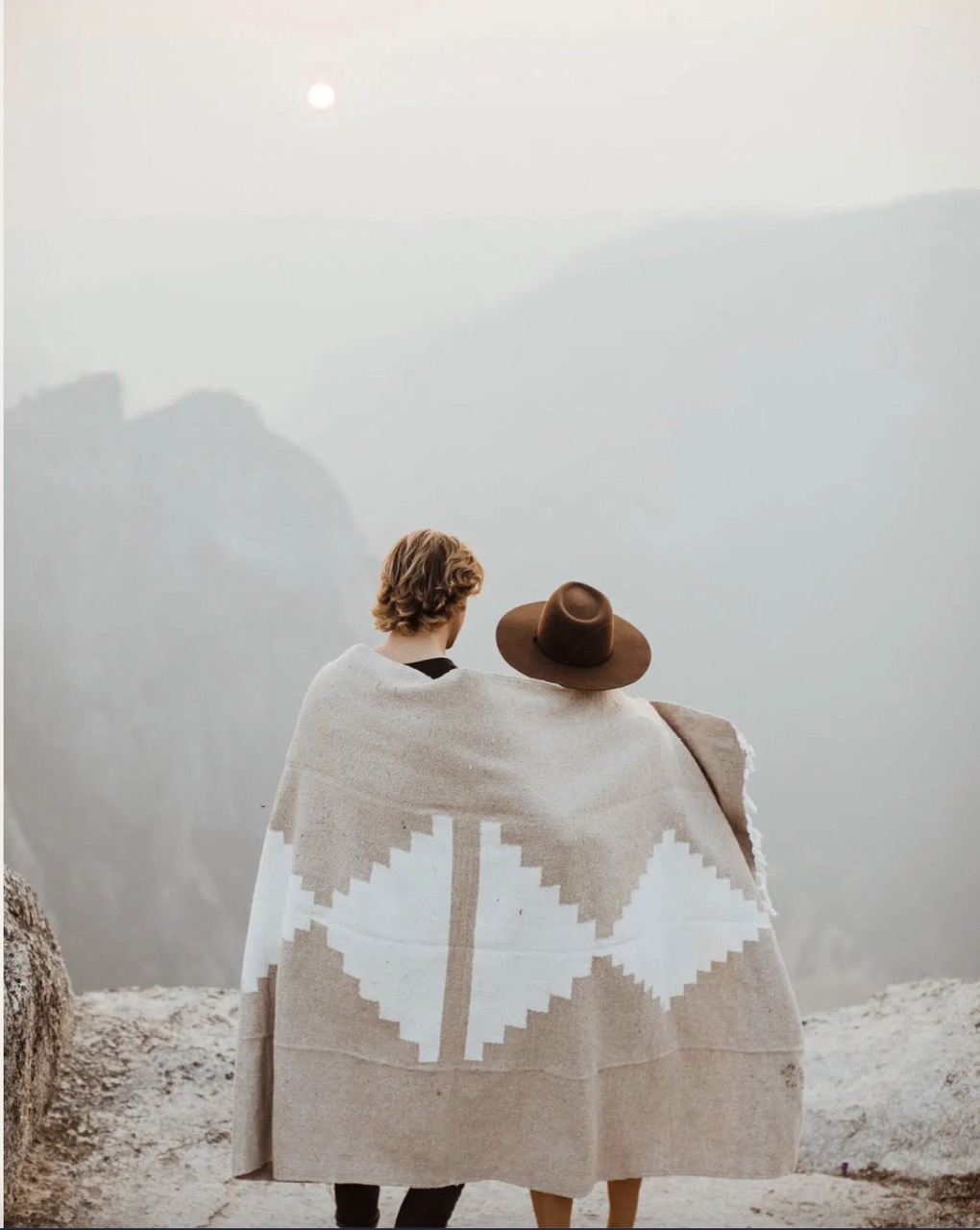 A woman in a hat and man stand in front of a mountain range with a woven beige blanket around them.