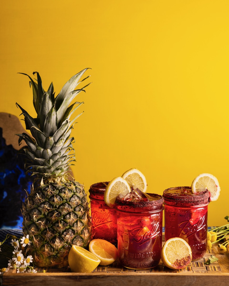Three glasses of red Juneteenth drinks featuring lemon wedges on the rim sit to the right of a pineapple in front of a yellow background.
