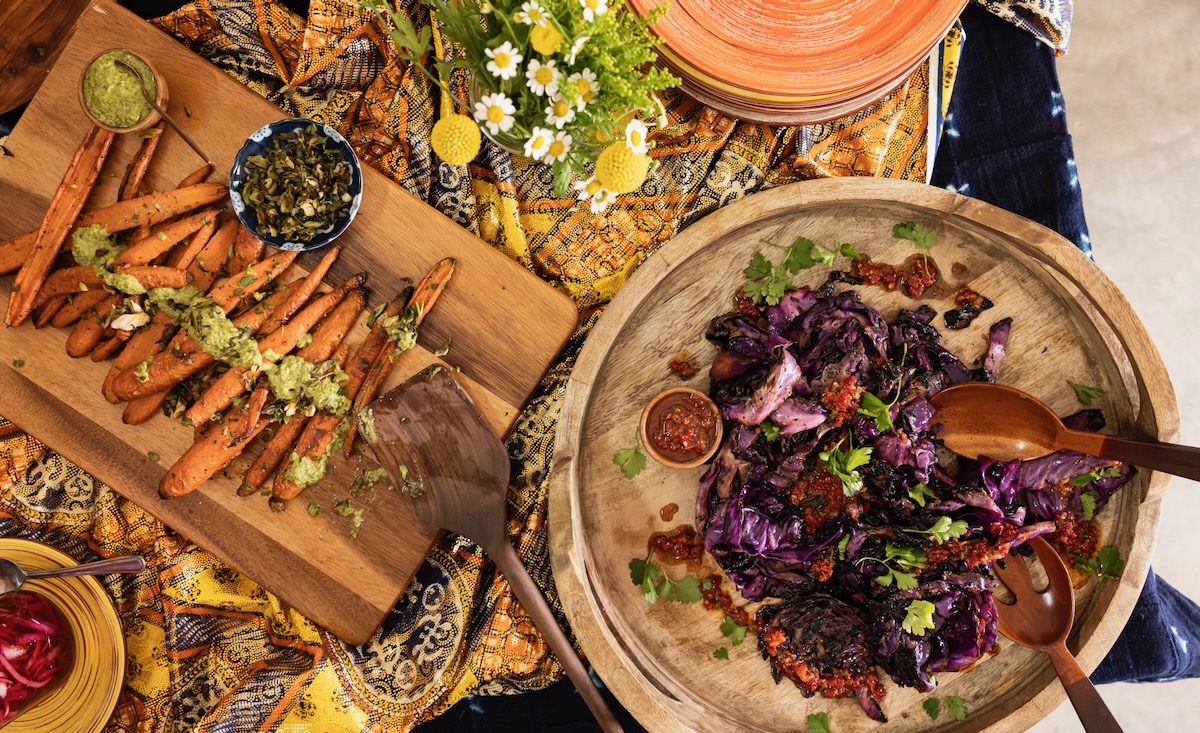 A wood cutting board with carrots sits to the left of a basket with charred red cabbage as a part of a Juneteenth picnic spread.