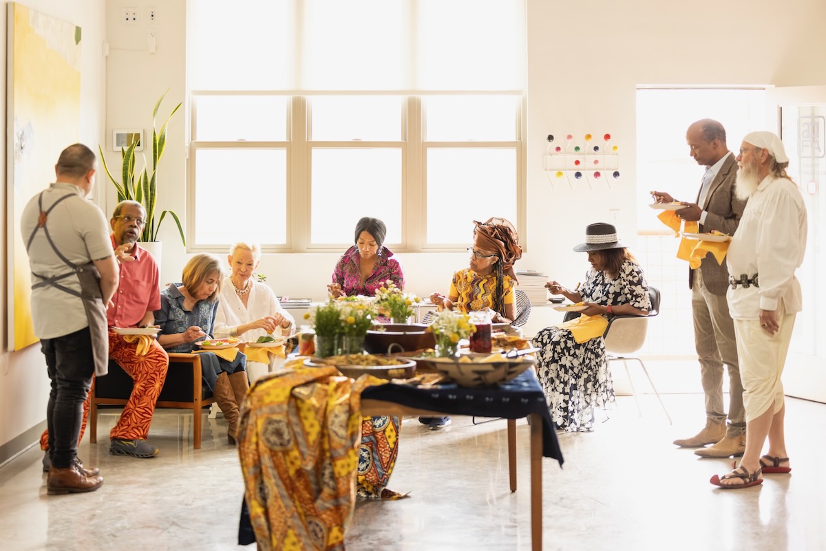 A group of Juneteenth event planners gather in a white room, plates in hand while enjoying picnic food.