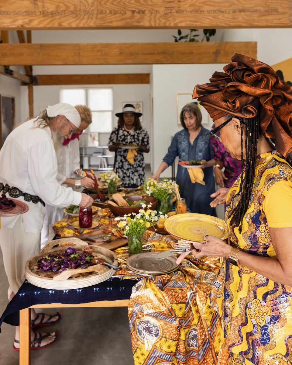 Various Juneteenth planners grab picnic food that's been spread around a wooden table with many colors of scarves decorated throughout.