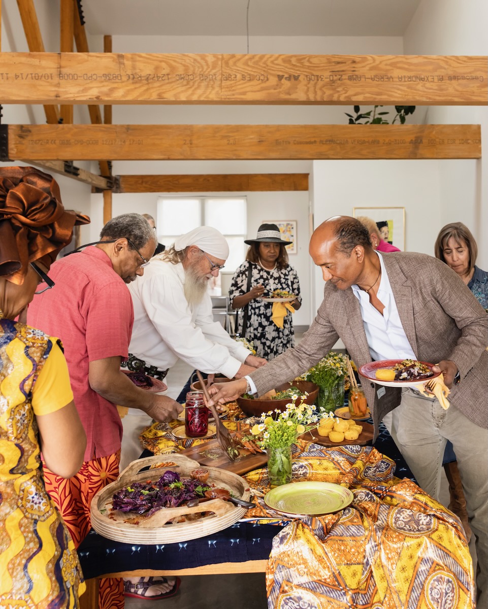 Various people grab food that's been spread around a wooden table with many colors of scarves decorated throughout.