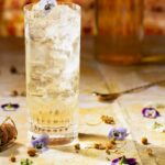 A clear glass with a spritzy cocktail inside sits on a brown table with violets and flowers scattered about next to a bar spoon.