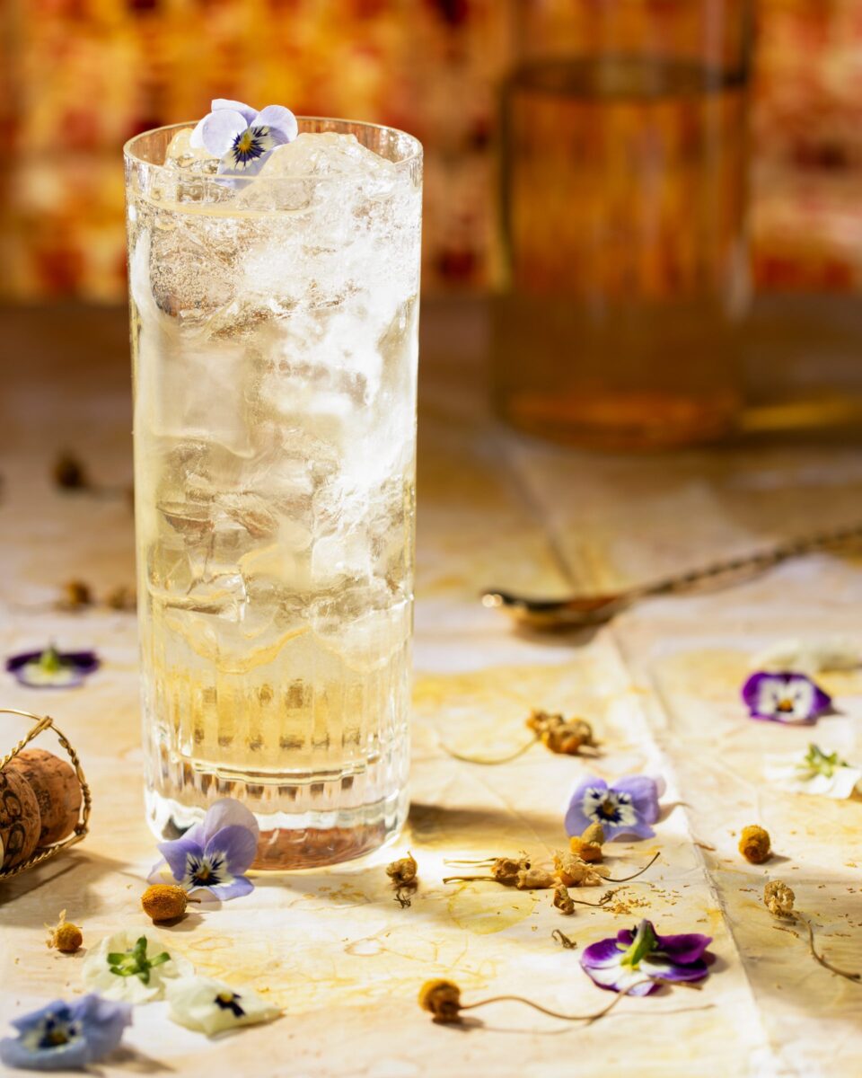 A clear glass with a spritzy cocktail inside sits on a brown table with violets and flowers scattered about next to a bar spoon.