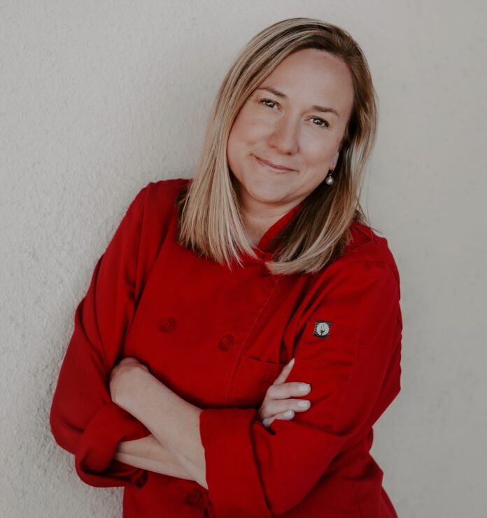 Laura Crucet stands in a red chef's jacket with her arms crossed in front of her chest and a smile on her face.