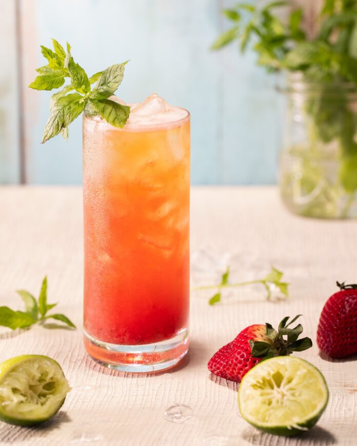 A tall collins glass holds a Min and Body summer cocktail recipe in a sunset gradient with a sprig of mint garnish and various strawberries and limes covering the table in front of the glass.