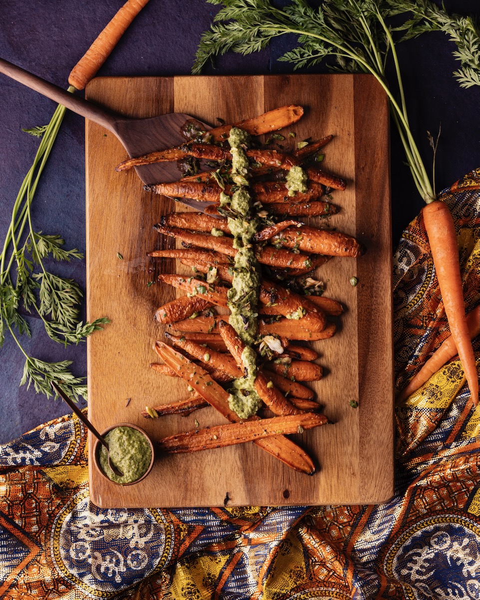 A wooden cutting board holds a line of oven-roasted sliced carrots with a walnut pesto lined down the center of the carrots.