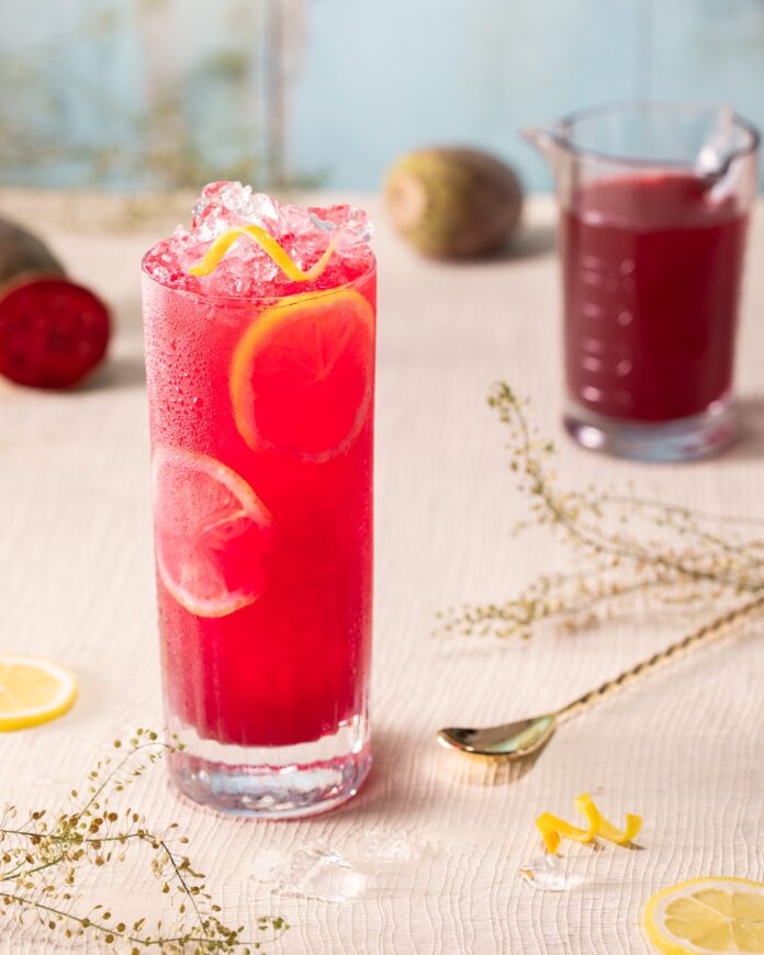 A glass of dark Pink Pony Club cocktail sits on a table with lemon slices in it on a white marble table top. A glass of dark purple prickly pear syrup sits in the background.