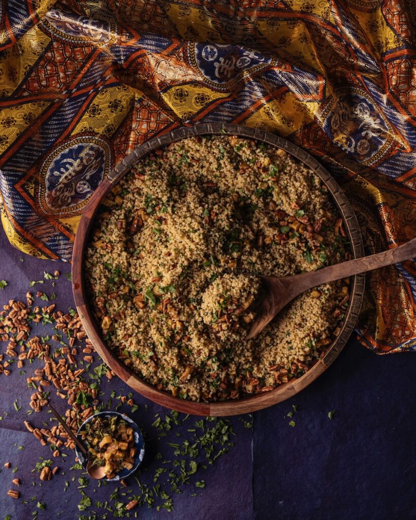 A brown bowl of a grain millet salad sits on top of a traditional cloth on a blue table as a spoon sits inside the grain bowl.