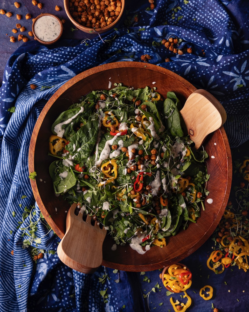 A big brown bowl holds a Spinach Salad with Blackened Chickpeas, banana peppers, and dressing with two wooden servers in the bowl as well, all against a blue tablecloth.
