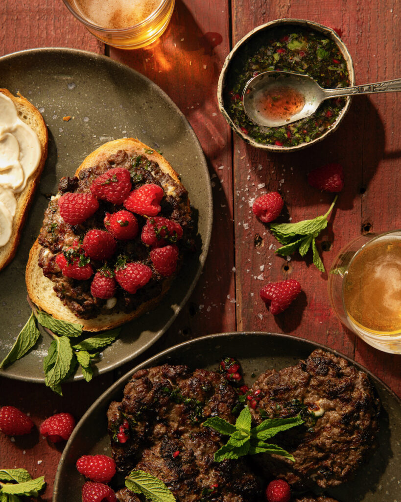 A lamb burger sits on a green plate topped with berries with stray berries and mint are scattered on the table.