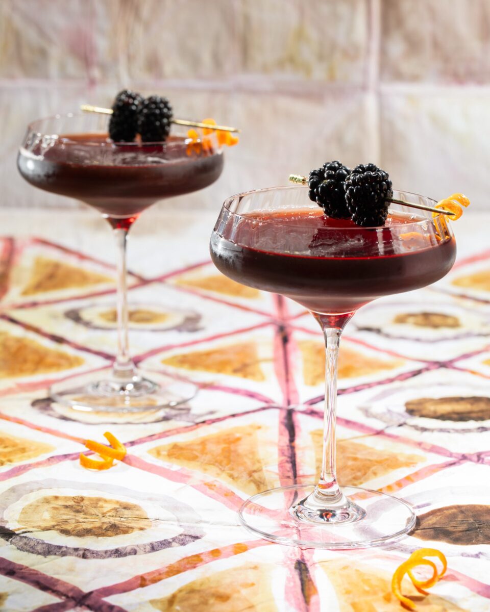 Two coupe glasses hold an Elixir of Love cocktail in a dark red color, garnished with two blackberries poked through a skewer as both glasses sit on a patterned table.