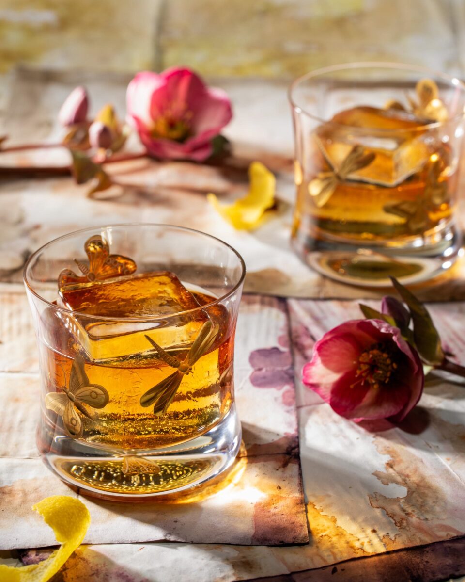 Two small cocktail glasses hold a brown cocktail with dragonflies on the outside of the glasses. Pink flowers are spread across the table.