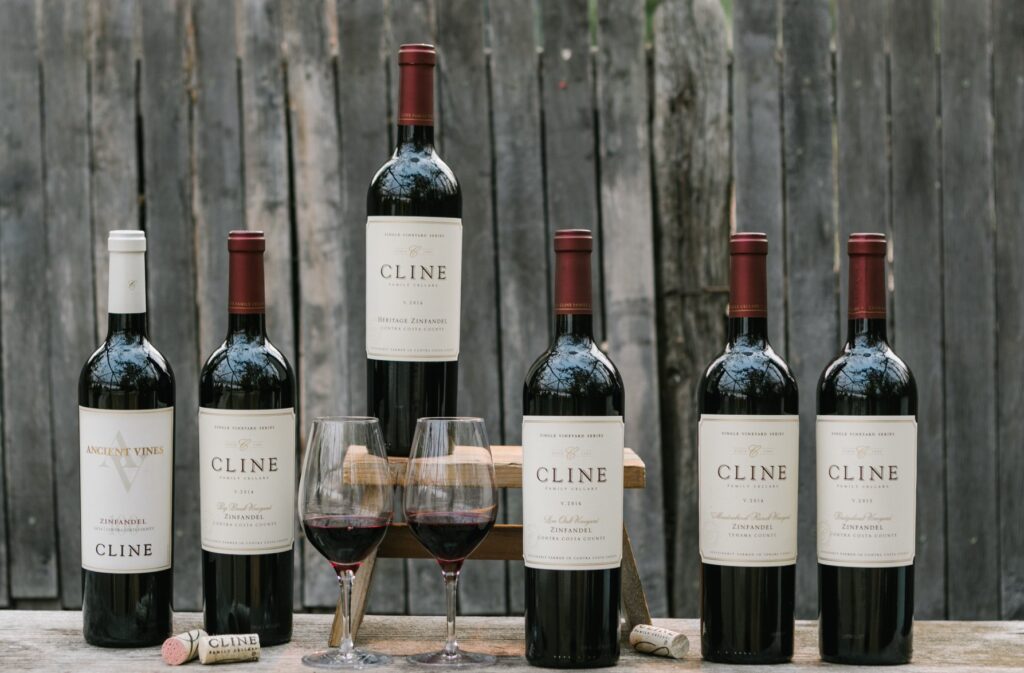 5 wines sit on a wood table behind a wood background with another bottle standing on a platform above the rest, featuring two glasses of wine below it.