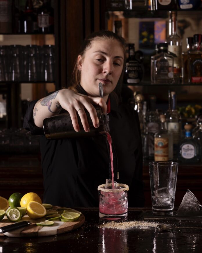 A bartender at La Posada pours a purple drink from high above intoa. rocks glass below.