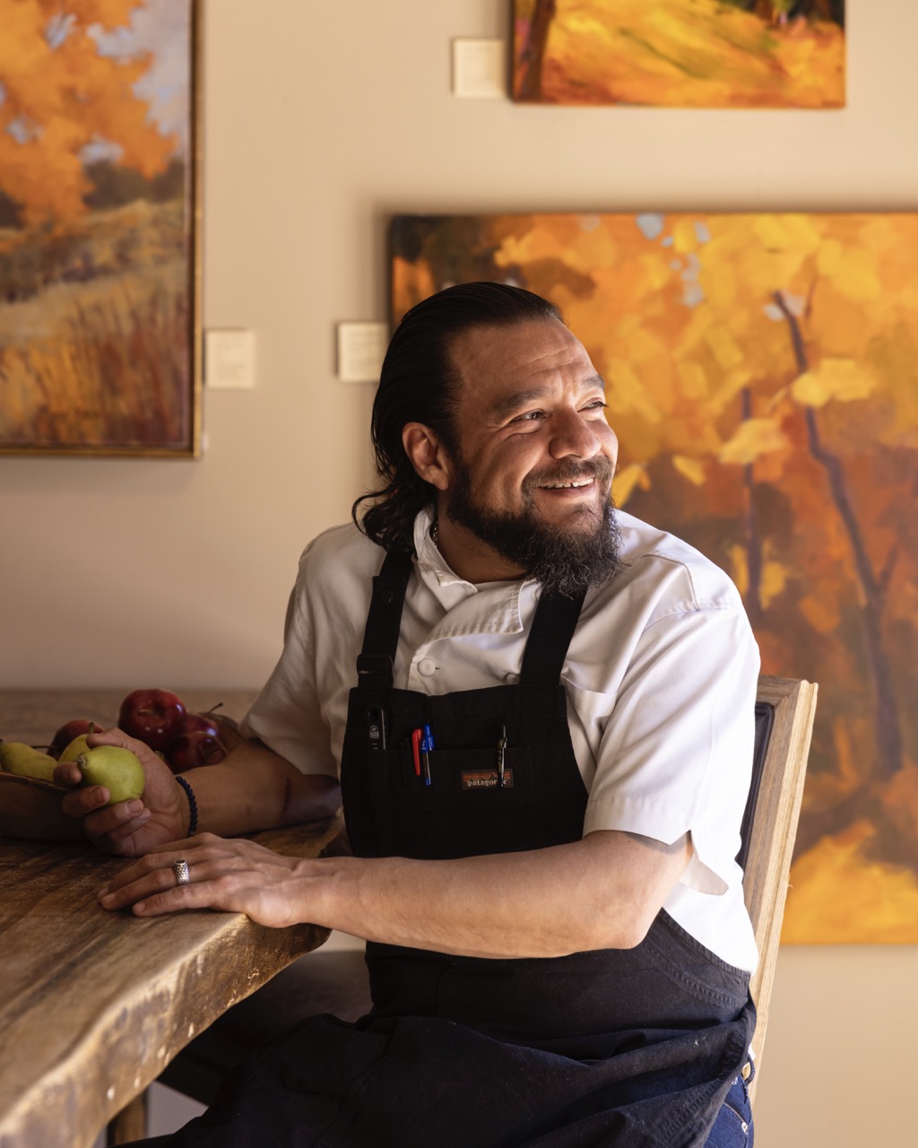 Chef Israel at La Posada sits at a bar in front of artwork in a chef's apron.