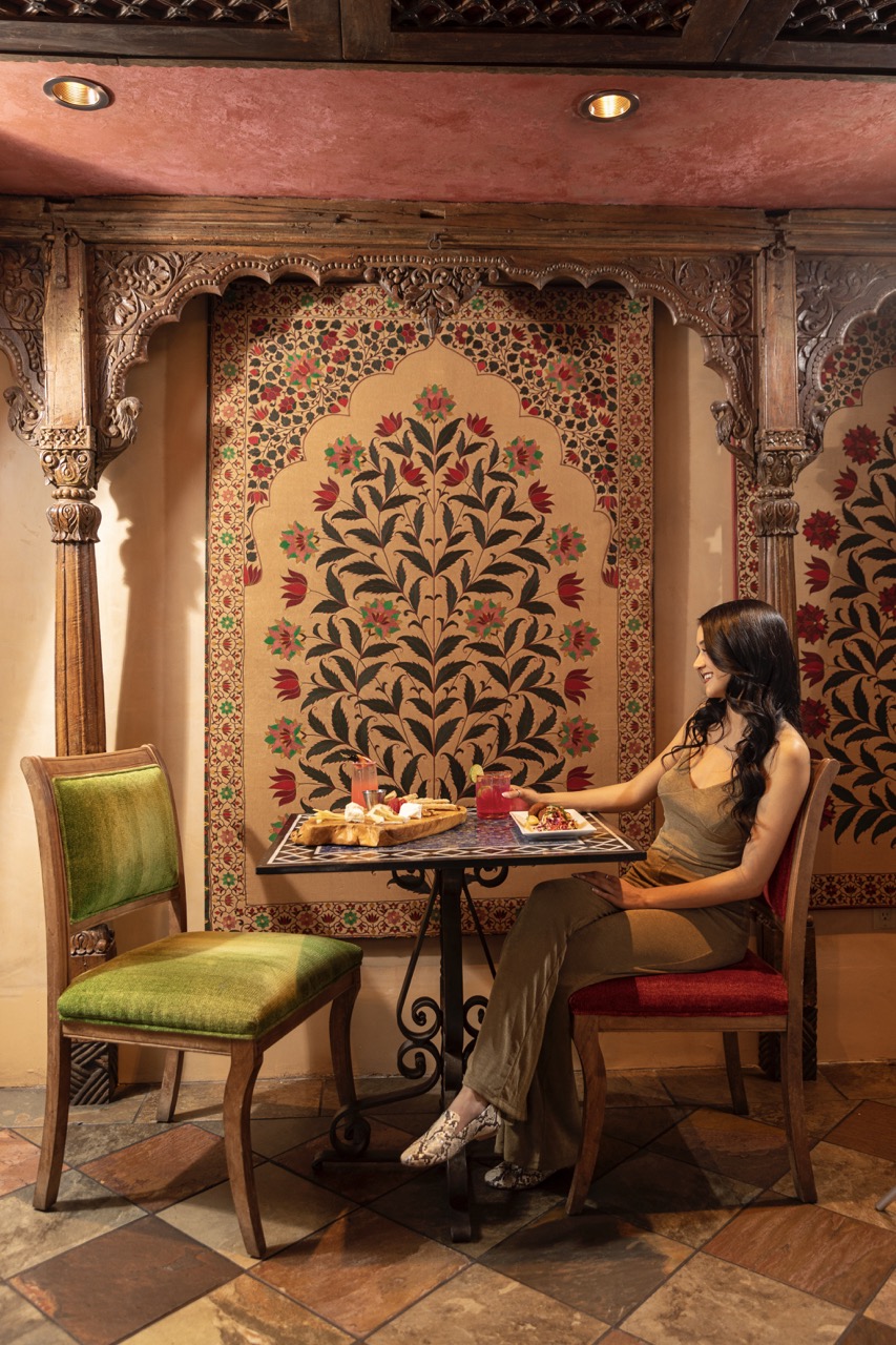 A woman sits in a red chair in front of a small table with amenities and a tapestry behind her. 