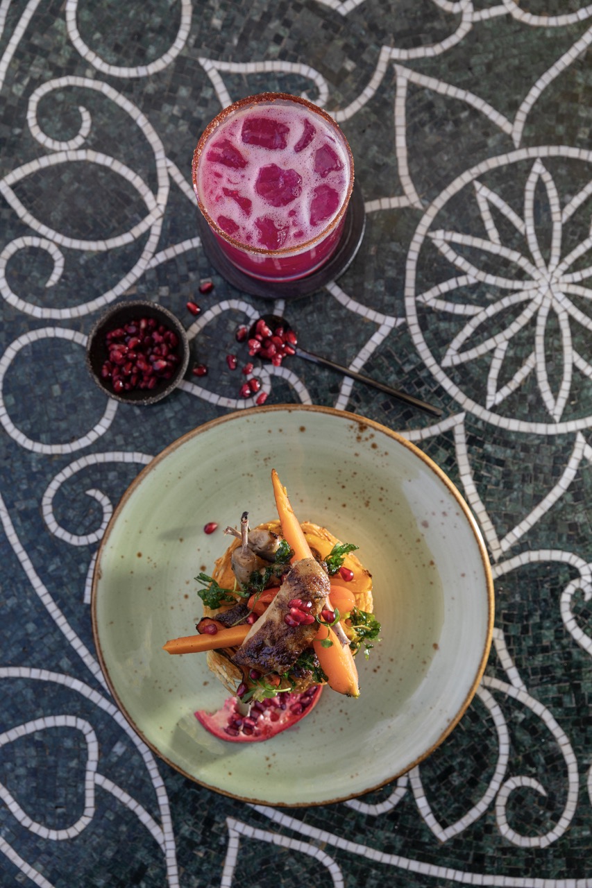 A greenish plate with food on top sits below a pink cocktail on a pattern table.