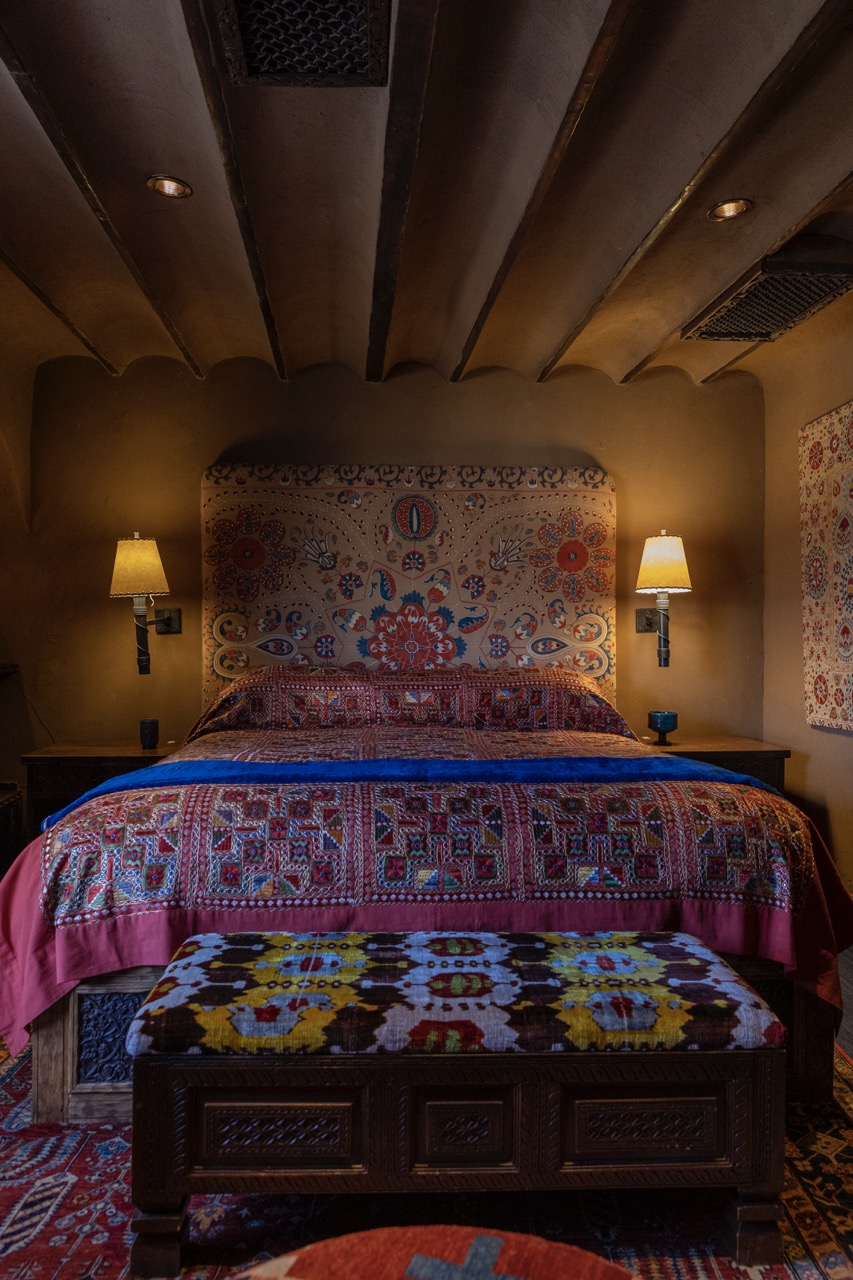 A bed sits in the center of a room with a pink and blue blanket and two lamps on the wall on either side of the bed. 