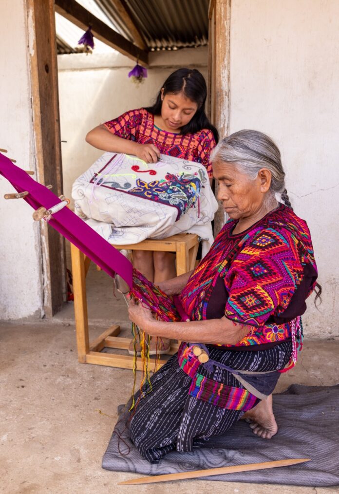 A woman with a grey ponytail weaves a rug while a young girl weaves a tapestry of her own.