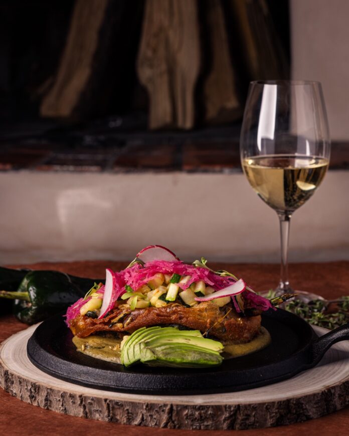 A black plate holds a fish dish topped with a slaw and with a wine glass to the right of the dish with a white wine.